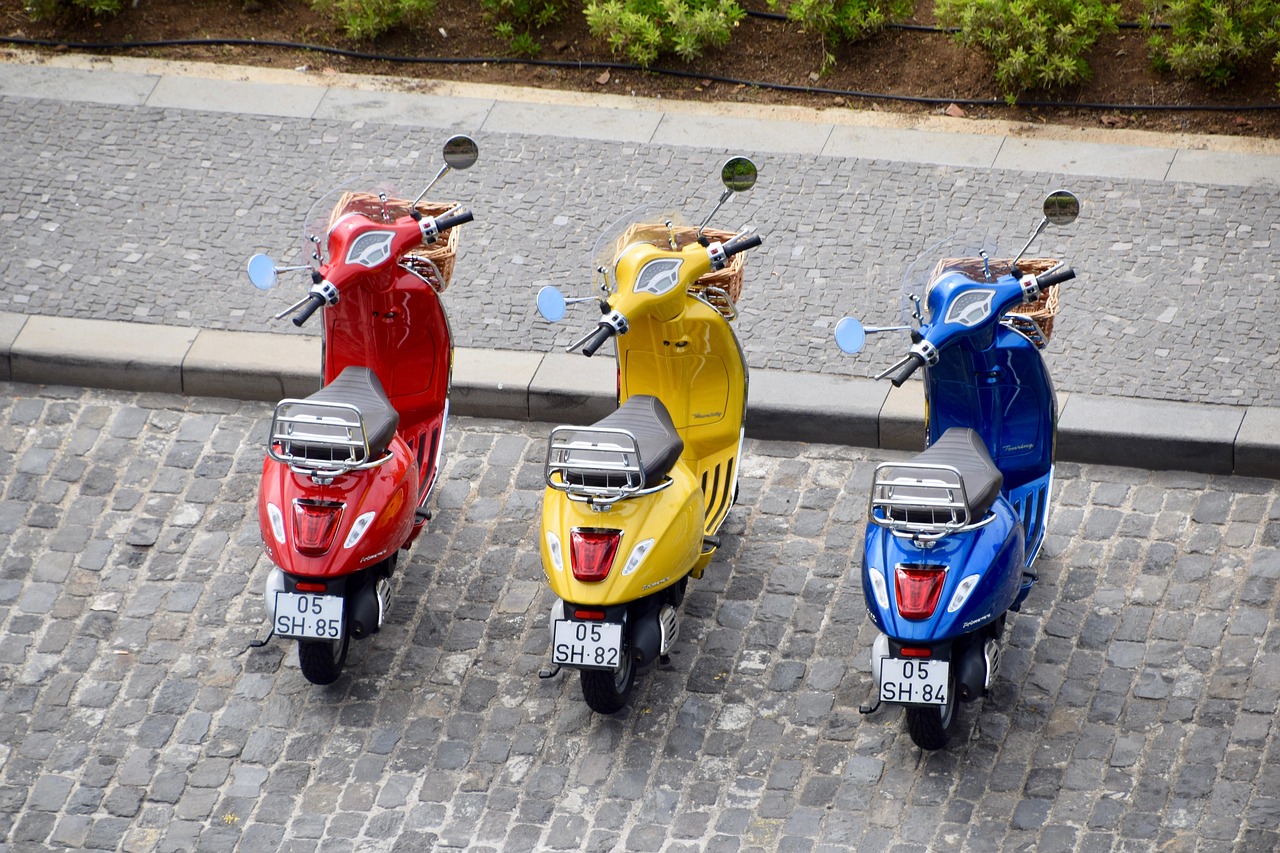 mopedy, kolobežky, vespa, piaggio, motocykle, bicykle, červená, žltá, modrá, tri, farebné, dláždené ulice, chodník, poznávacie značky, držadlá, sedadlá, svetlá, zrkadlá, kolesá, šedé ulice, šedá zrkadlo, šedá bicykli