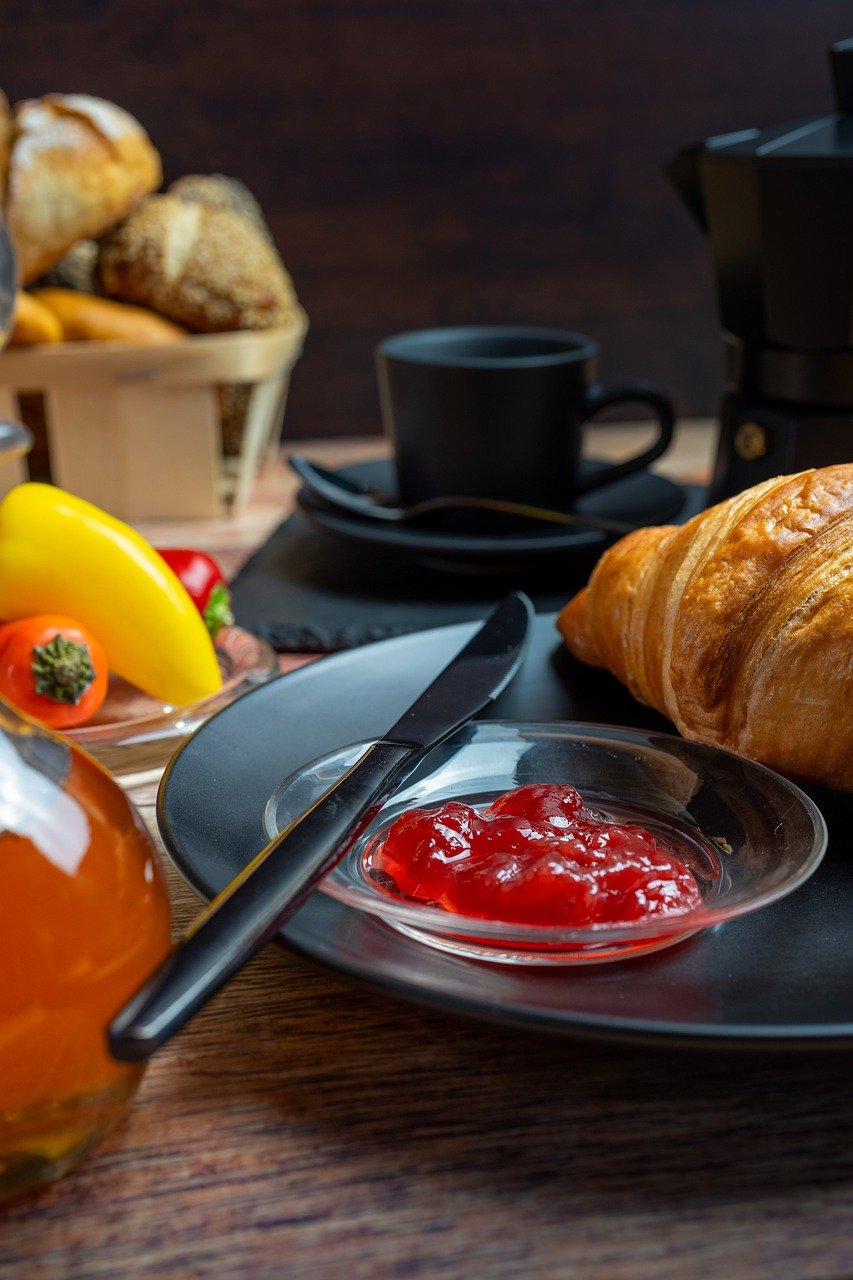 breakfast, croissant, jam, honey, coffee machine, coffee pot, black dishes, bread basket, bun, paprika, tomatoes, rustic table, baked goods, morning meal, mediterranean, comfortable, food scene, enjoyment, café atmosphere